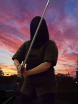 Gillian Dean in full fencing mask and padded jacket with German longsword against a sunset sky.